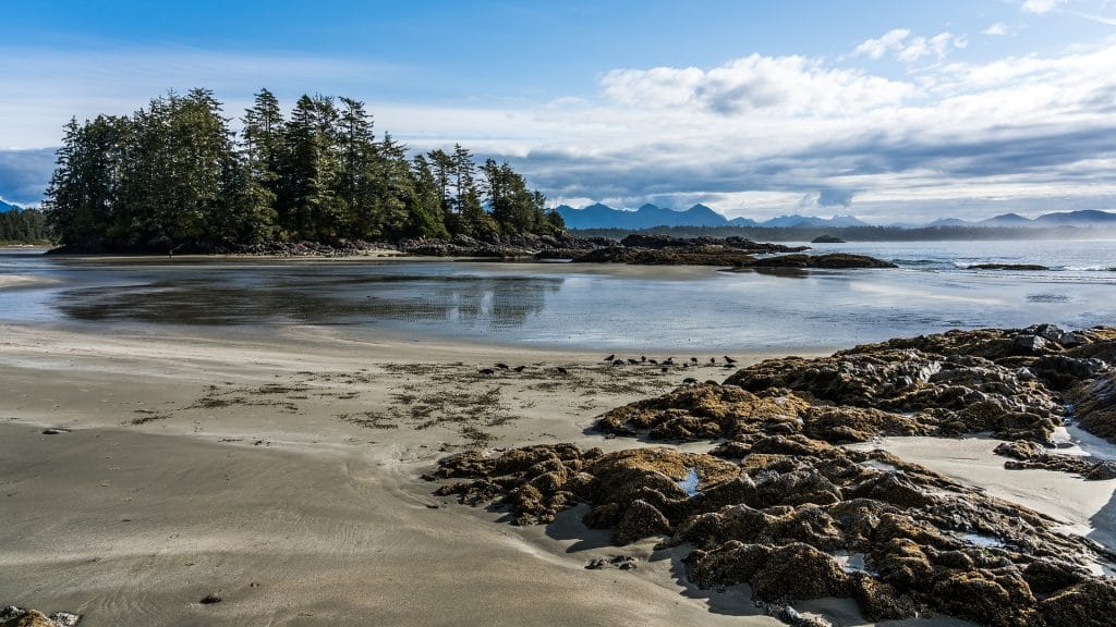 Rocky coastline with sandy beach, shallow water, and forested island under a partly cloudy sky; distant mountains visible in the background—one of the best places to visit in BC for scenic coastal views.