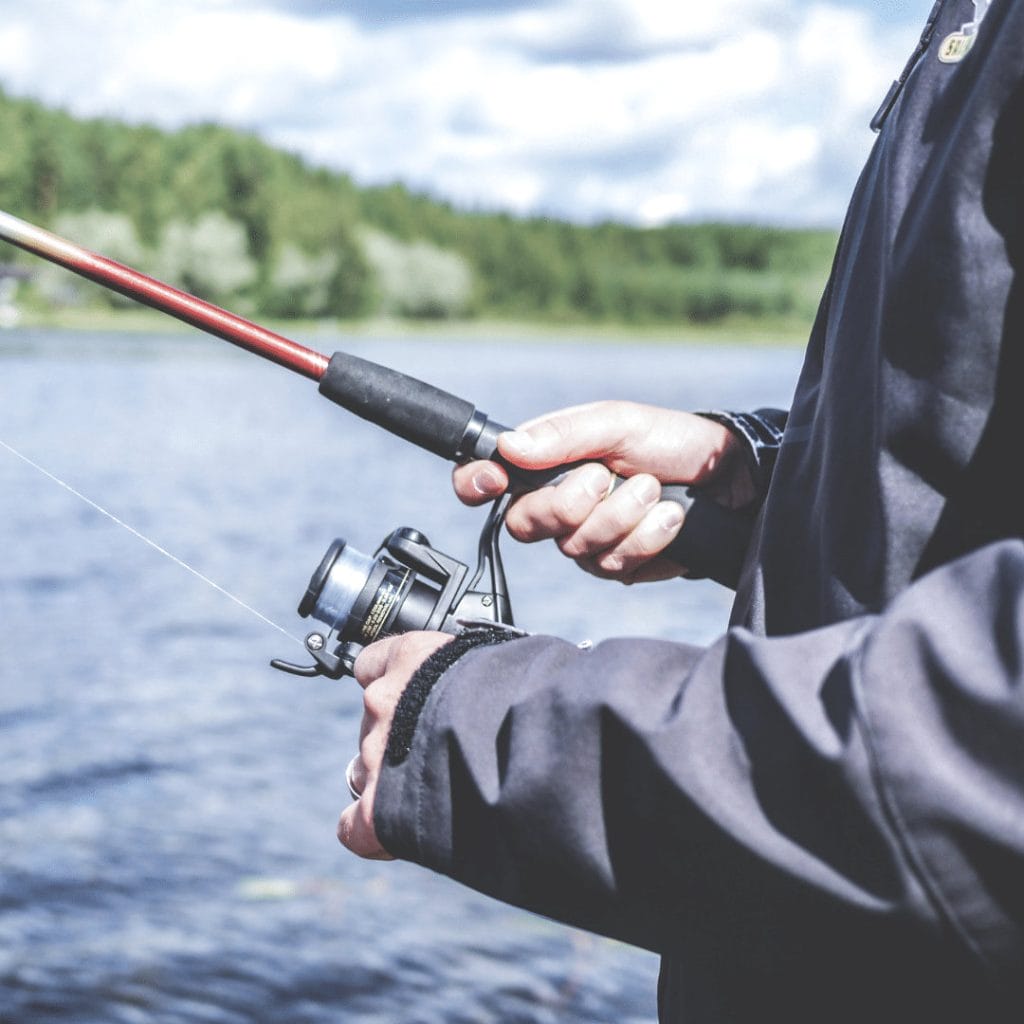 Person holding a fishing rod and reel by a body of water, wearing a dark jacket, with trees and sky in the background—capturing one of the favorite things to do in Ucluelet.