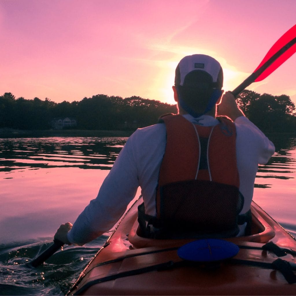 Kayaking on a calm lake at sunset is one of the best things to do in Ucluelet, where you can glide past trees and houses in the distance, wearing a white shirt, life vest, and cap.