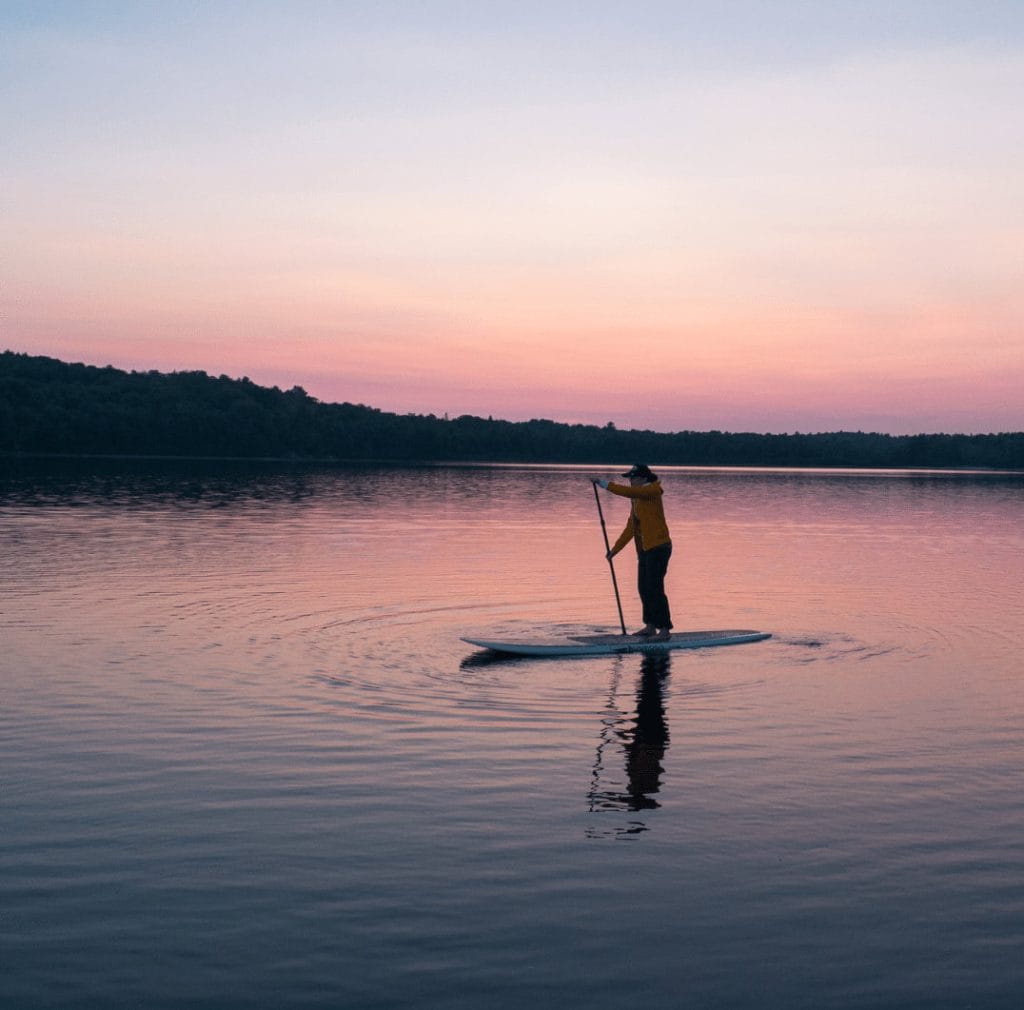 A person paddleboards on a calm lake at sunset, with pink and purple hues in the sky and water—a serene scene showcasing one of the best things to do in Ucluelet, with a tree-lined shore visible in the distance.