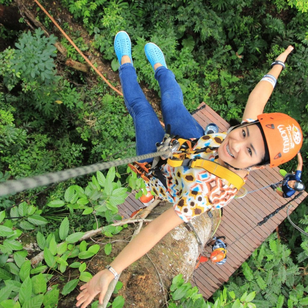 A person in a helmet and harness is zip-lining above a forest, smiling and extending their arms outward, with a wooden platform and other people visible below—one of the exciting things to do in Ucluelet.