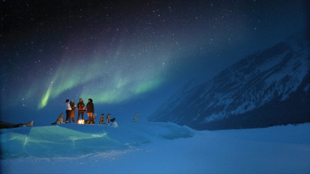 A group of people and dogs stand by a campfire on snowy terrain under the northern lights near Banff, with a mountain in the background at night.