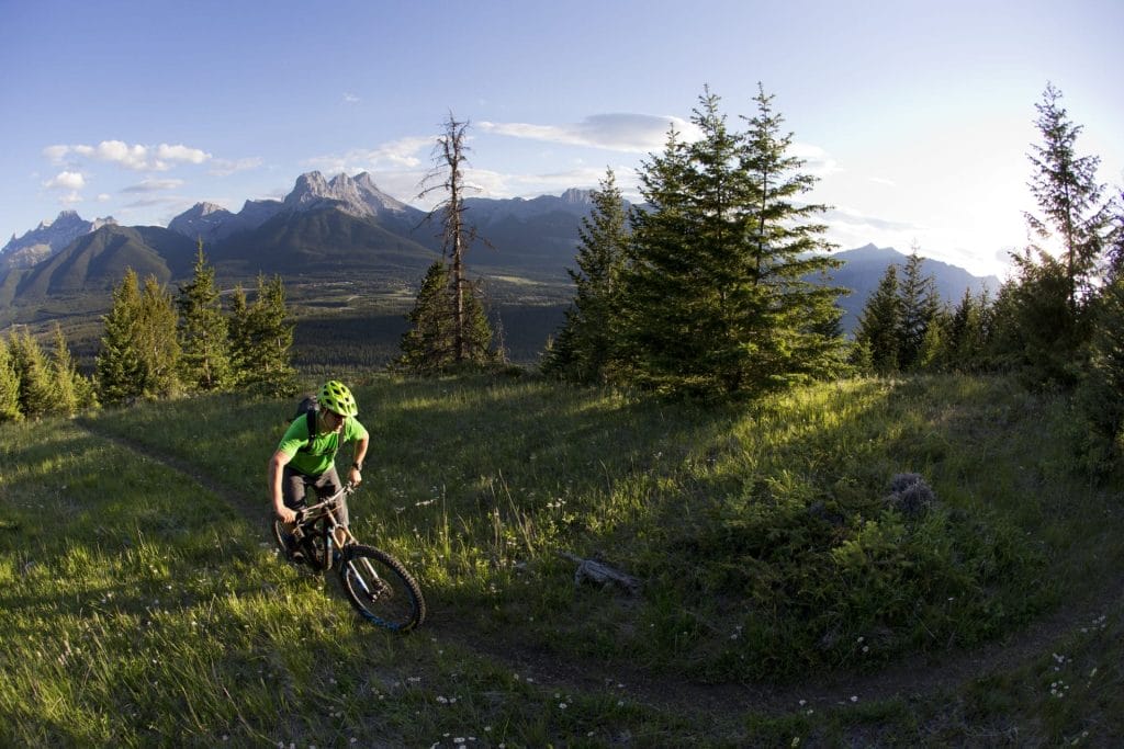 A cyclist wearing a green helmet and shirt rides a mountain bike on a grassy trail near Canmore, with mountains and trees in the background.