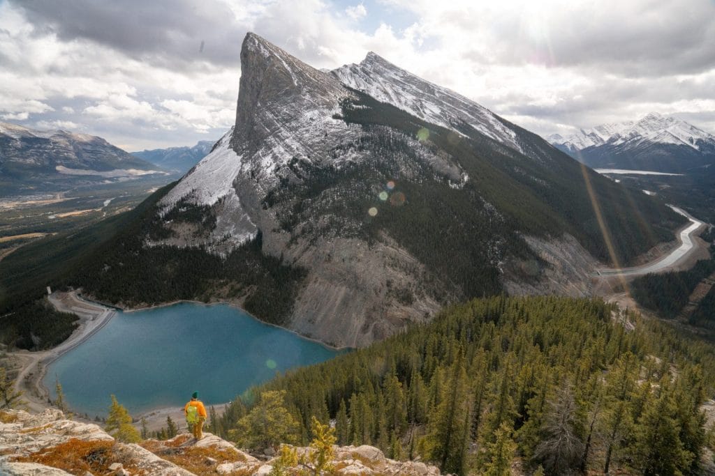 A person in an orange jacket stands on a rocky slope overlooking a turquoise lake near Banff, dense forest, and a steep, snow-dusted mountain peak under a cloudy sky.