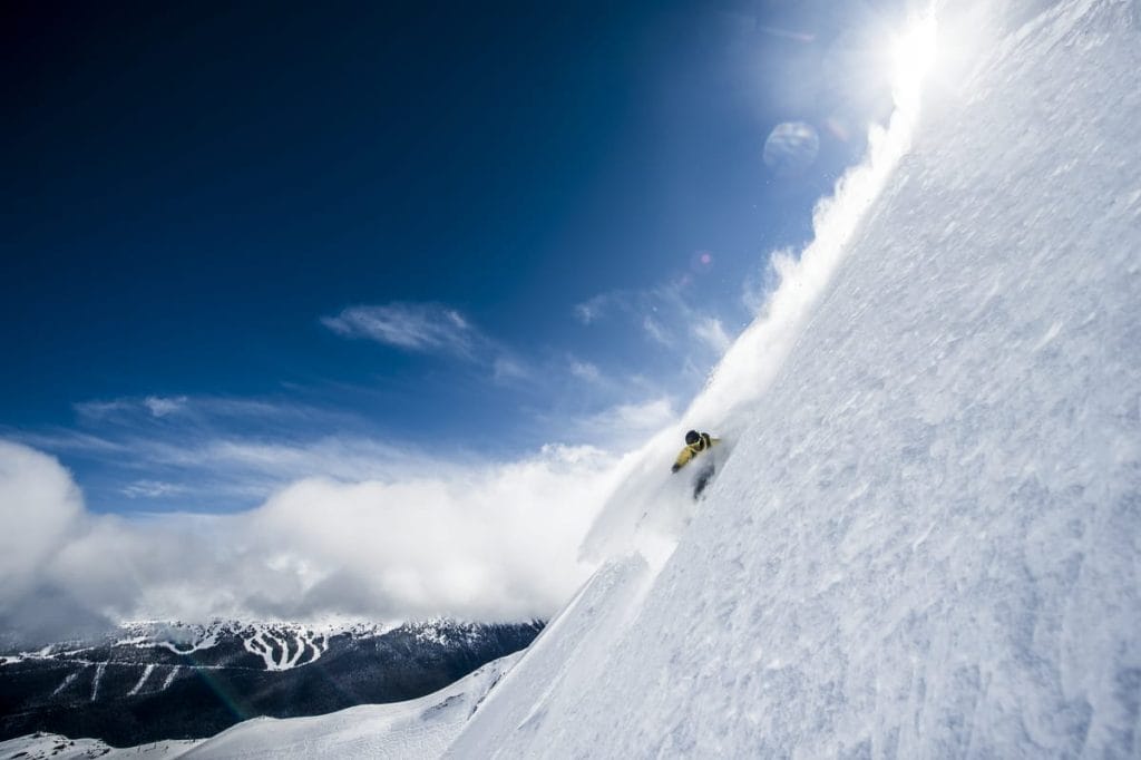 A skier in a yellow jacket descends a steep, snowy slope with powder spraying, under a clear blue sky and bright sunlight—true adventure awaits along the Sea to Sky Brewery Guide.