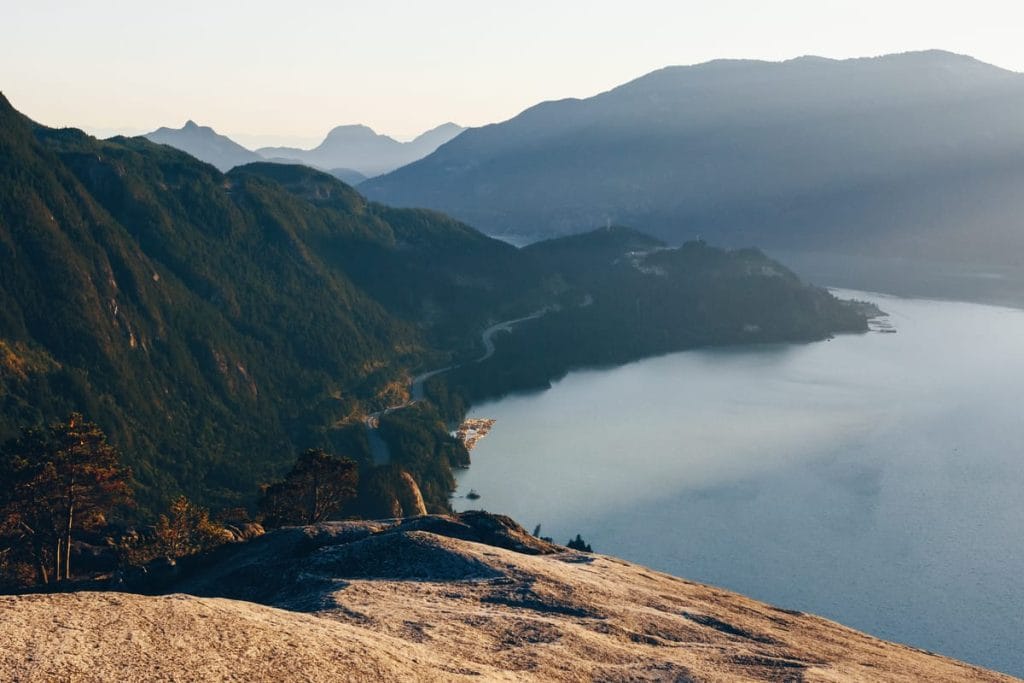 View of a coastal landscape with forested mountains, a winding road, and a calm body of water under a clear sky at sunset—perfect scenery to pair with your Sea to Sky Brewery Guide adventure.