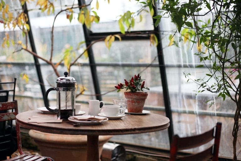 A round wooden table with a French press coffee maker, two cups and saucers, and a potted plant, set by large windows with plants visible outside—perfect for cozy mornings in short-term rentals.