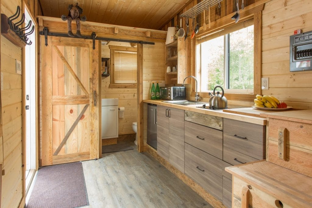 A cozy kitchen with wood walls and cabinets, a large window, sink, microwave, kettle, bananas on the counter, and a sliding barn door to the bathroom—perfectly organized according to the Airbnb Host Checklist for guest comfort.