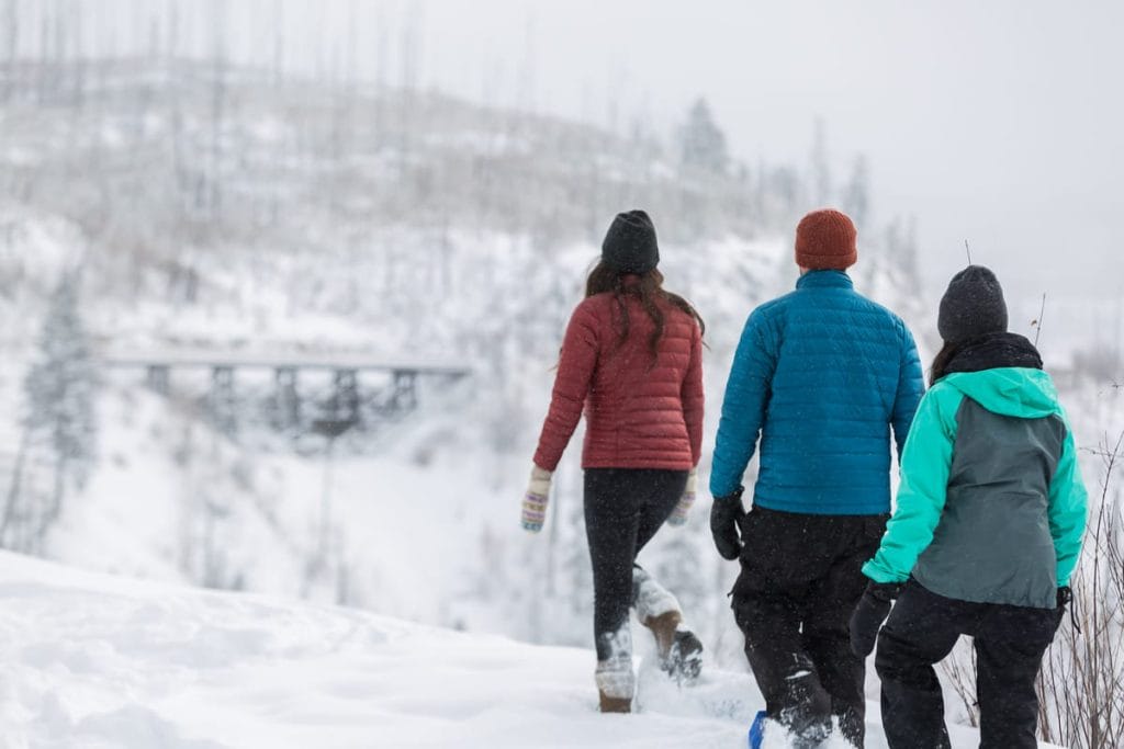 Three people wearing winter jackets and hats walk through a snowy landscape, exploring snow-covered trees and a bridge in the background—a perfect example of winter things to do in Kelowna.