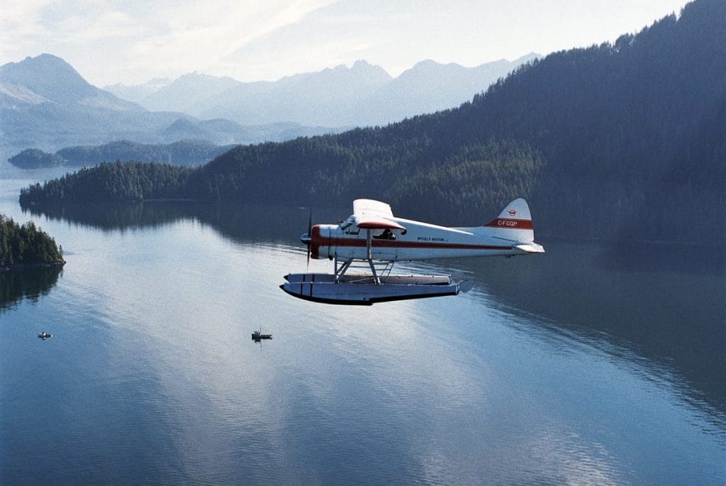 A seaplane flies over a calm lake surrounded by forested mountains, with several small boats below—capturing the adventurous spirit of things to do in Tofino.