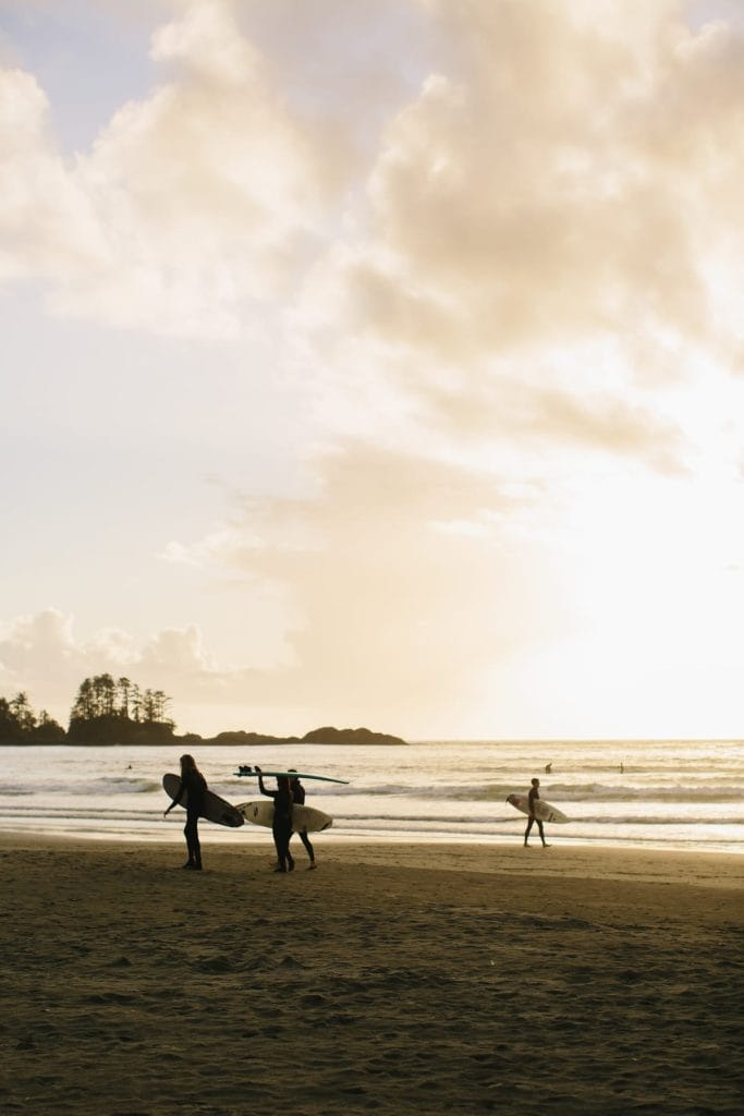 Three surfers with surfboards walk along a sandy beach at sunset, with waves in the background and a cloudy sky overhead—one of the top things to do in Tofino for adventure seekers and ocean lovers.