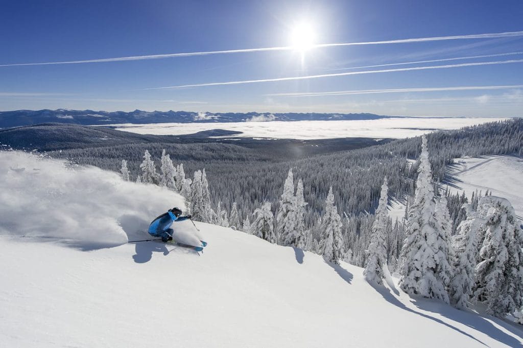 A skier in blue gear descends a snowy slope, kicking up powder, with snow-covered trees, mountains, and a bright sun in the background—one of the exhilarating things to do in Kelowna.