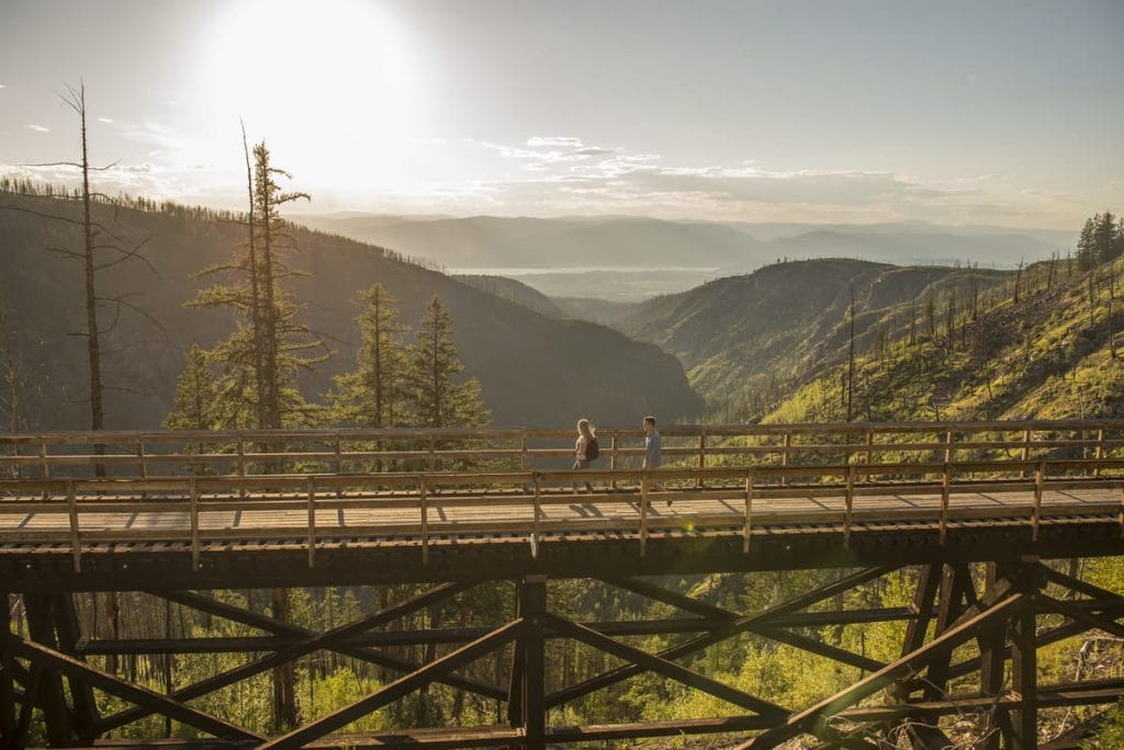 Two people walk on a wooden bridge with a scenic view of mountains, trees, and the bright sun—a perfect moment among the many memorable things to do in Kelowna.