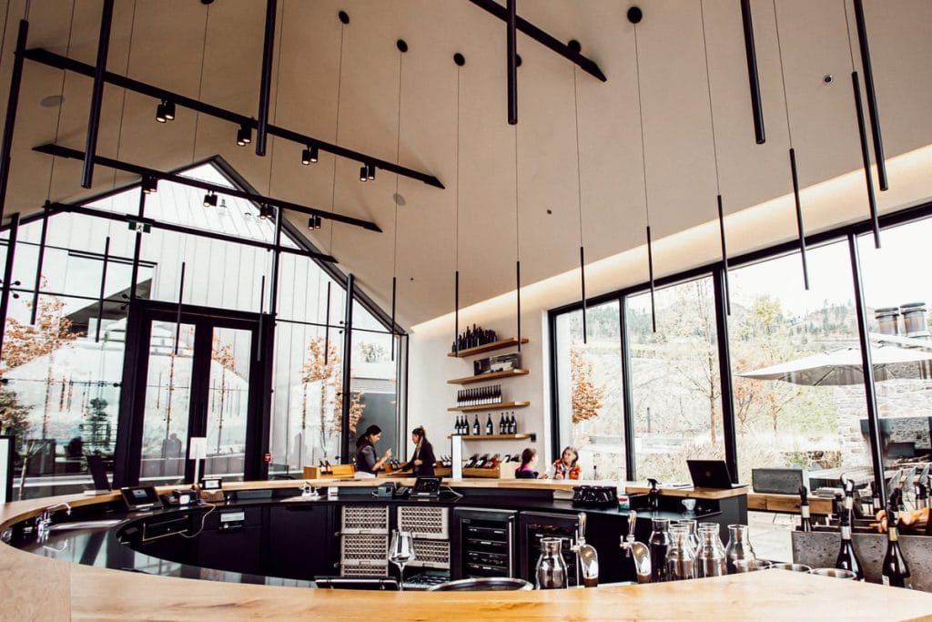Modern bar interior with high ceilings, hanging lights, and large windows—an ideal spot to unwind after exploring Things to Do in Kelowna. People sit at the counter, with shelves of bottles and glassware visible behind the bar.