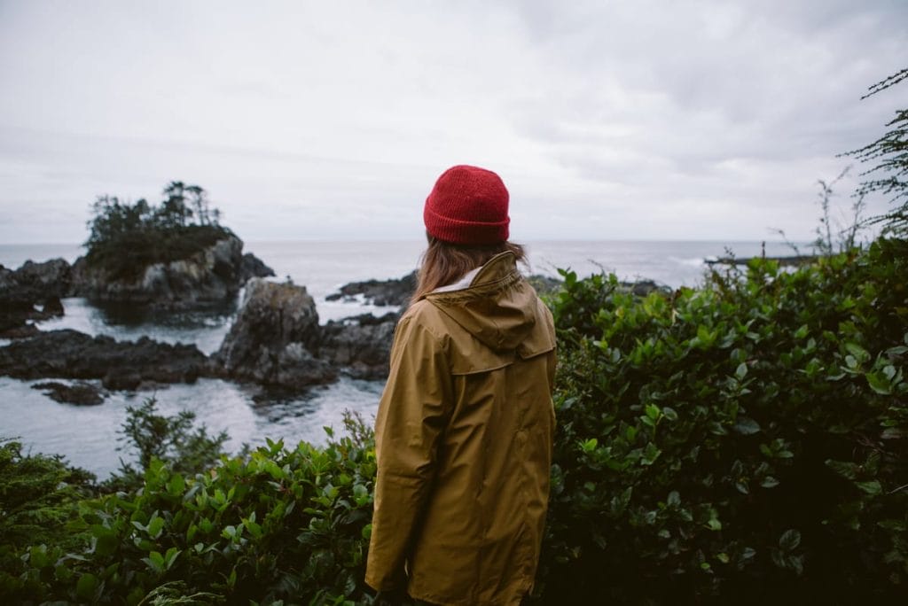 A person in a yellow rain jacket and red beanie stands by green bushes, looking out at a rocky coastline under a cloudy sky—one of the classic things to do in Tofino.