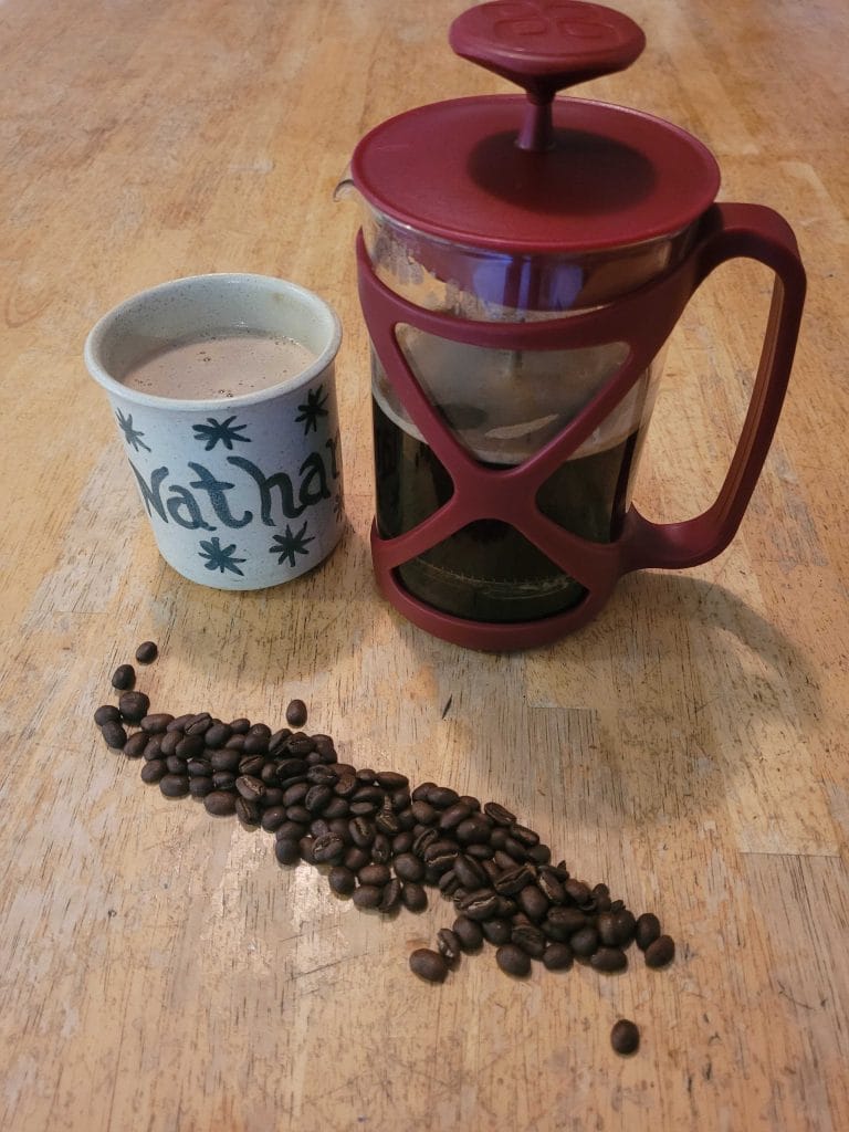 A French press with coffee, a personalized mug, and a small pile of coffee beans are arranged on a wooden table—perfect essentials for short-term rentals or anyone seeking the best coffee makers for their morning brew.