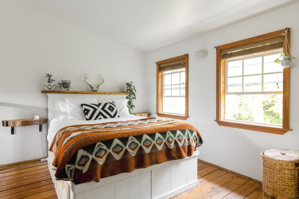 A bright bedroom showcases Vacation Rental Design with a double bed, geometric-patterned blanket, wooden floors, two windows, and minimalist decor including plants and antlers on a shelf.