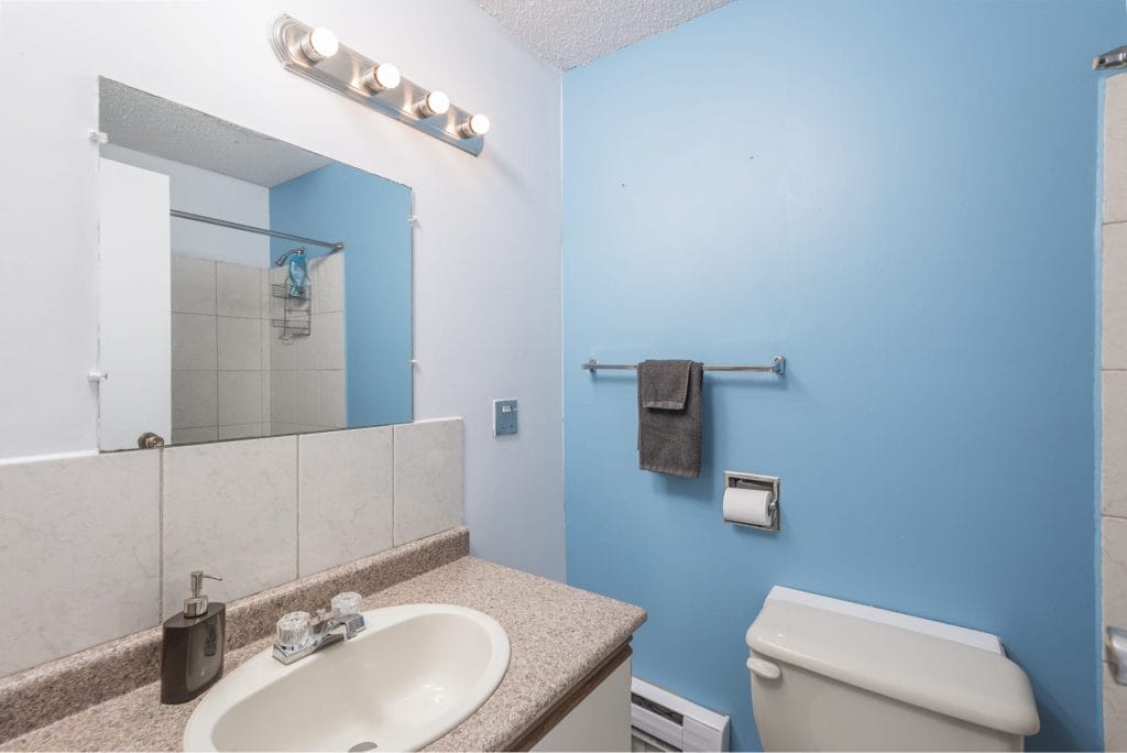Small bathroom with a blue wall, white sink, beige countertop, wall-mounted mirror, toilet, gray towel on a rack, and overhead light fixture—perfectly styled for modern vacation rental design.