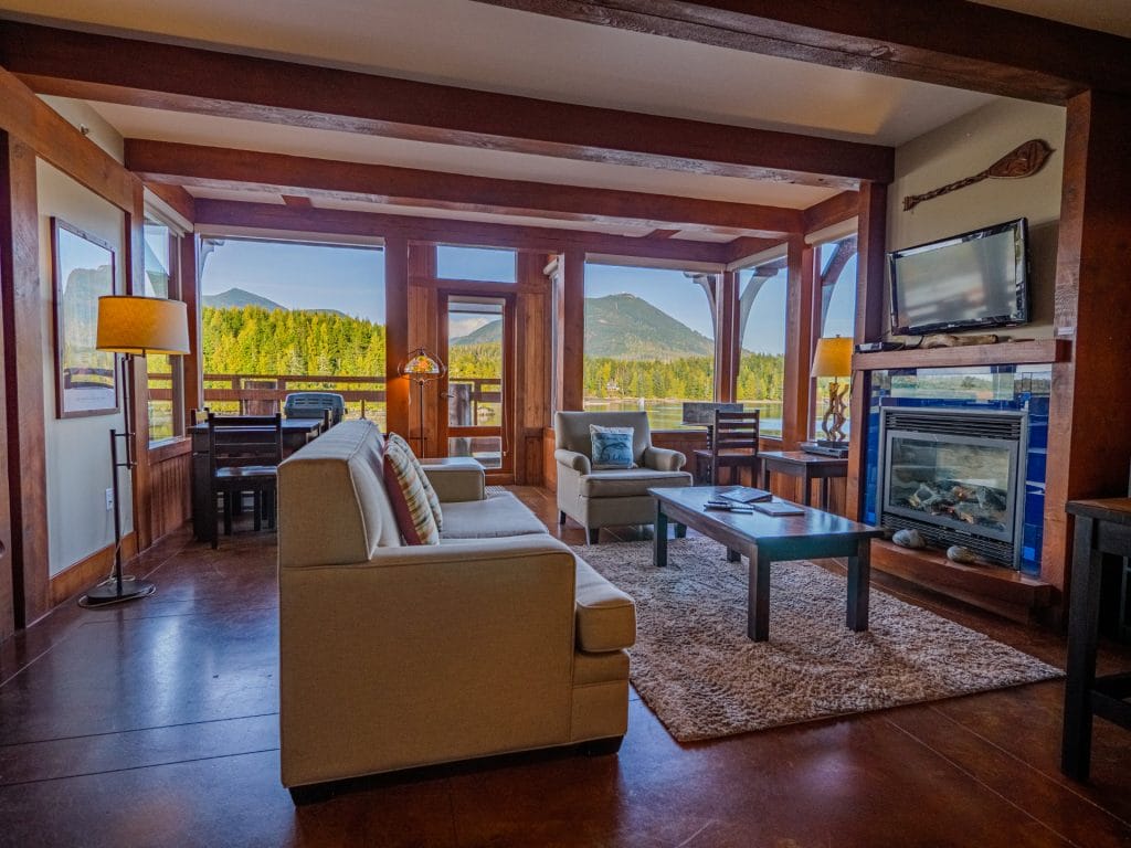 A cozy living room with beige sofas, a coffee table, a fireplace, a mounted TV, and large windows overlooking mountains and pine trees—perfectly crafted with Vacation Rental Design in mind.
