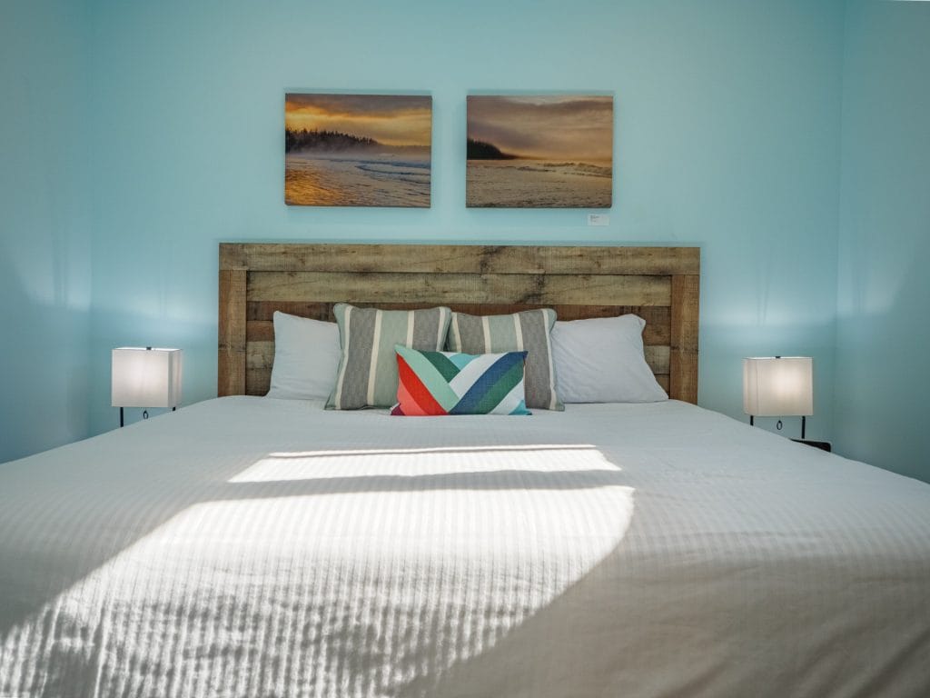 A neatly made bed with white linens, colorful accent pillow, two bedside lamps, and two landscape paintings on a light blue wall showcases thoughtful Vacation Rental Design.