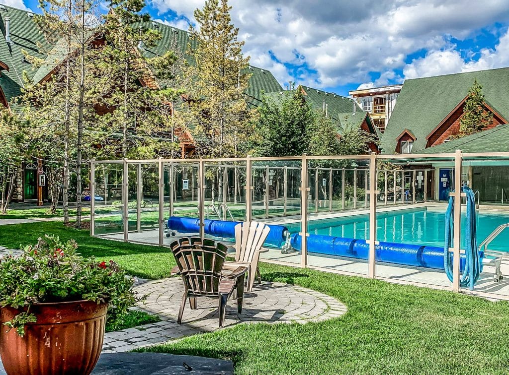 Outdoor swimming pool enclosed by a glass fence, surrounded by green grass and wooden cabins; patio chairs and a potted plant in the foreground showcase inviting vacation rental design.
