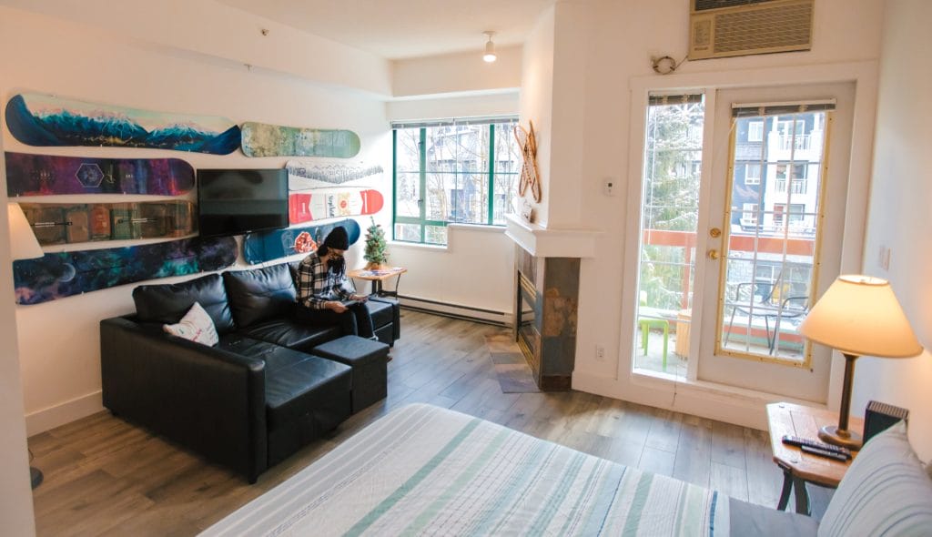 A small modern studio apartment with a bed, black couch, TV, and snowboard-themed wall art, showcases Vacation Rental Design with large windows and a glass door leading to a balcony.