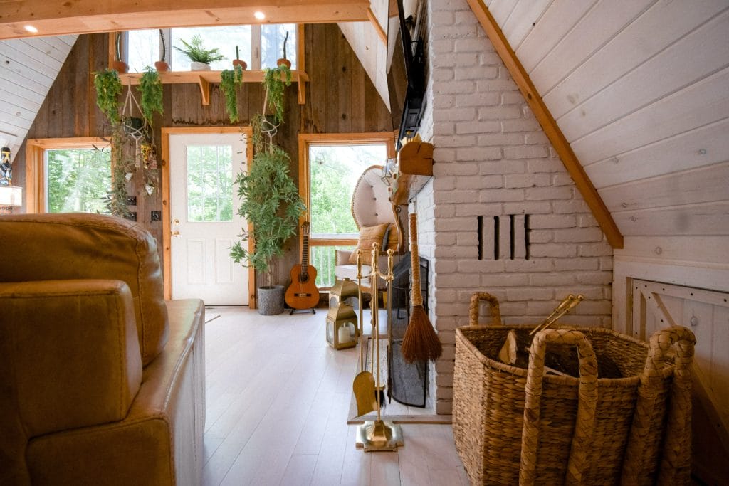 Cozy A-frame cabin interior showcases thoughtful Vacation Rental Design with wooden walls, a white brick fireplace, wicker baskets, fireplace tools, potted plants, and a guitar near the windowed door.
