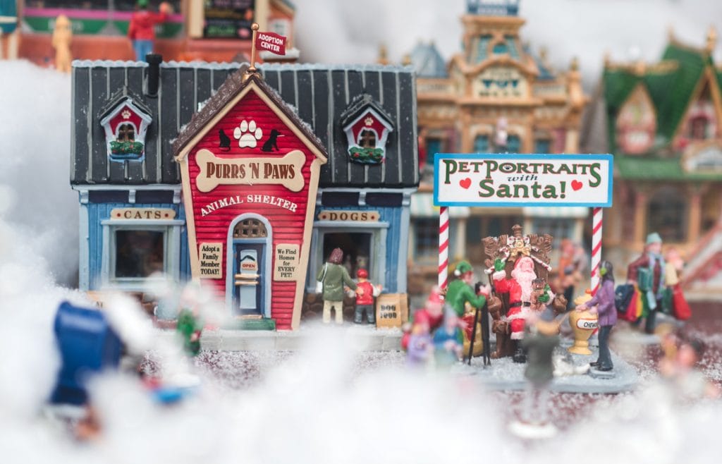 A miniature holiday village scene inspired by christmas markets in vancouver, with an animal shelter, people, and a "Pet Portraits with Santa!" stand featuring Santa Claus and pets.