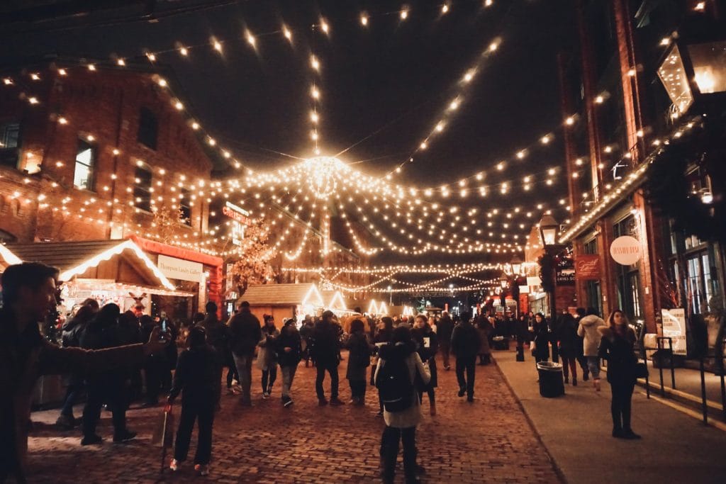 People walk along a cobblestone street at night under strings of lights, surrounded by market stalls and brick buildings—capturing the festive charm of Christmas markets in Vancouver.