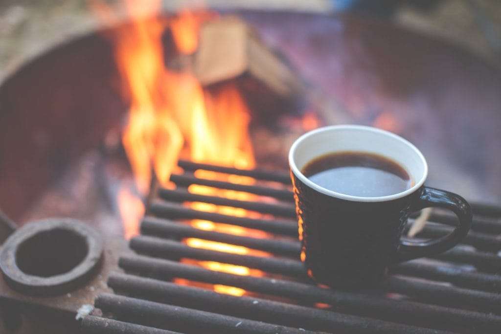 A black mug filled with coffee sits on a metal grill over a campfire, flames and logs burning in the background—showing how to brew coffee while camping and savor the outdoors.