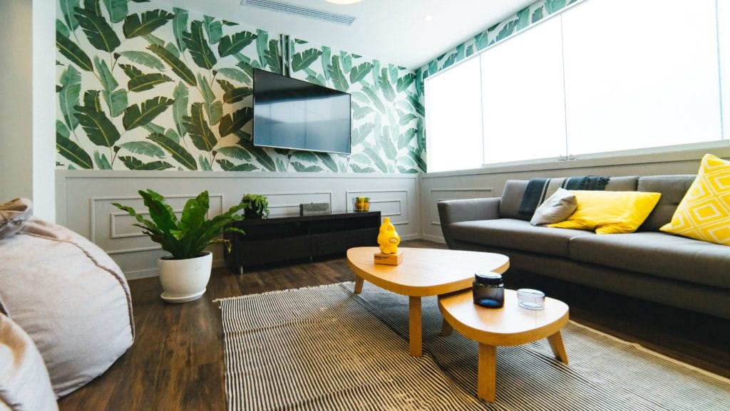 Modern living room in an eco-friendly Airbnb with a leafy wallpaper accent wall, mounted TV, gray sofa with yellow pillows, coffee table, potted plant, and large window letting in natural light.