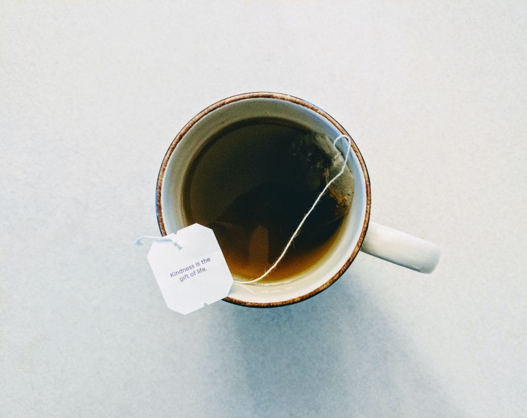 A white mug filled with tea sits on a light surface, with a tea bag and tag reading, "Kindness is the gift of life." Perfect for moments when you’re learning how to brew coffee while camping or simply enjoying some quiet time.