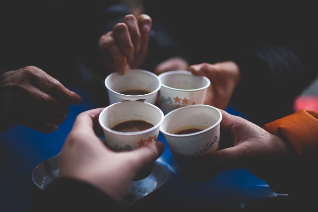 Four hands holding paper cups filled with a hot beverage, raised together in a casual toast—perfect for sharing stories about how to brew coffee while camping.
