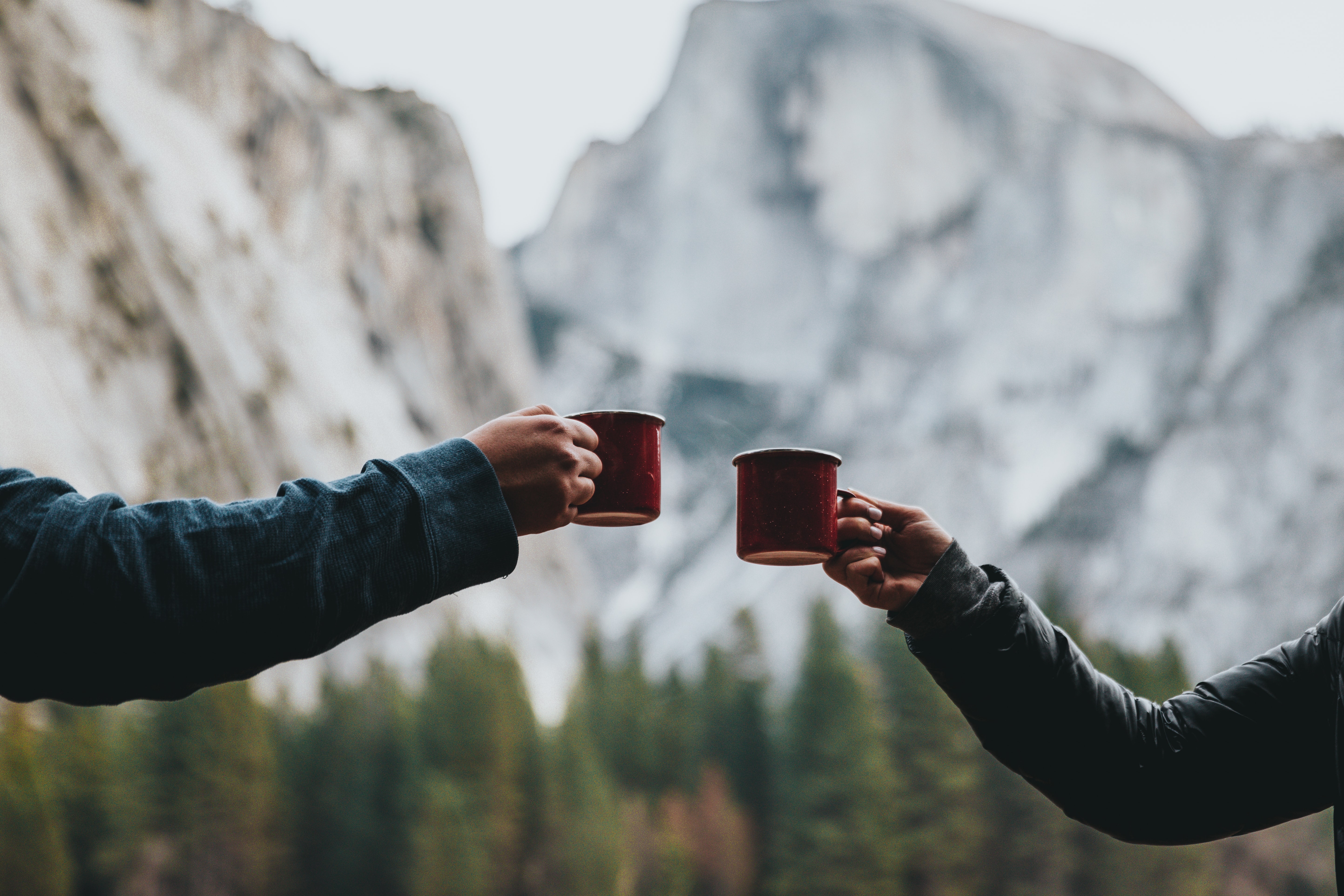 Two people toast with red mugs, celebrating their adventure in the wild. A forest and a rocky mountain rise behind them—capturing the perfect moment of learning how to brew coffee while camping in nature.