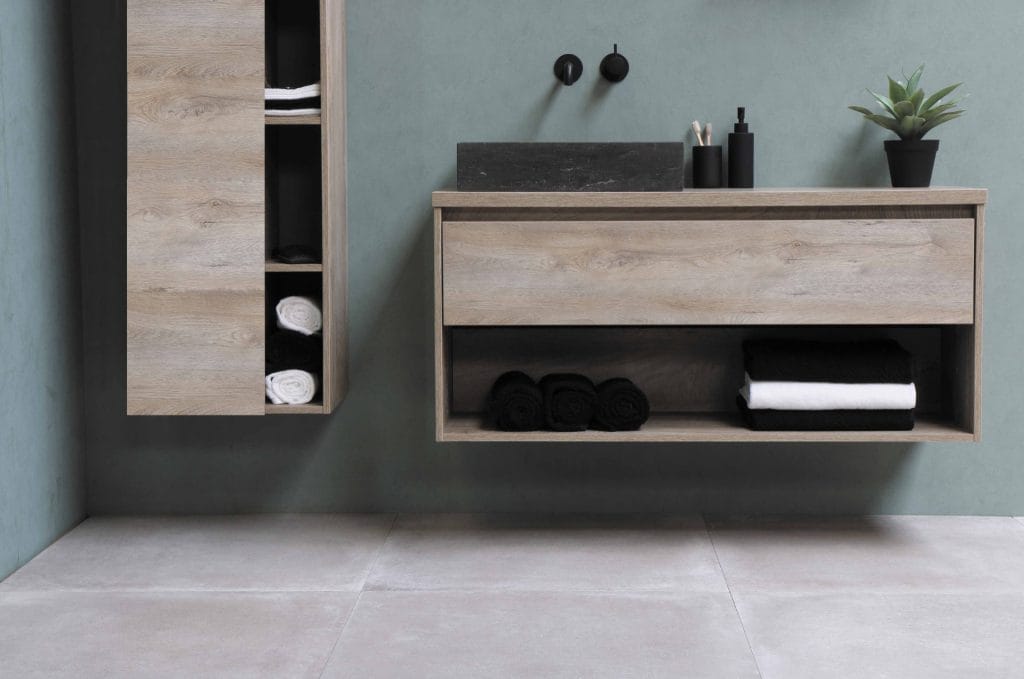 Minimalist bathroom in an eco-friendly Airbnb with light wood cabinets, rolled towels, a black sink with wall-mounted faucet, and a small potted plant on a green wall.