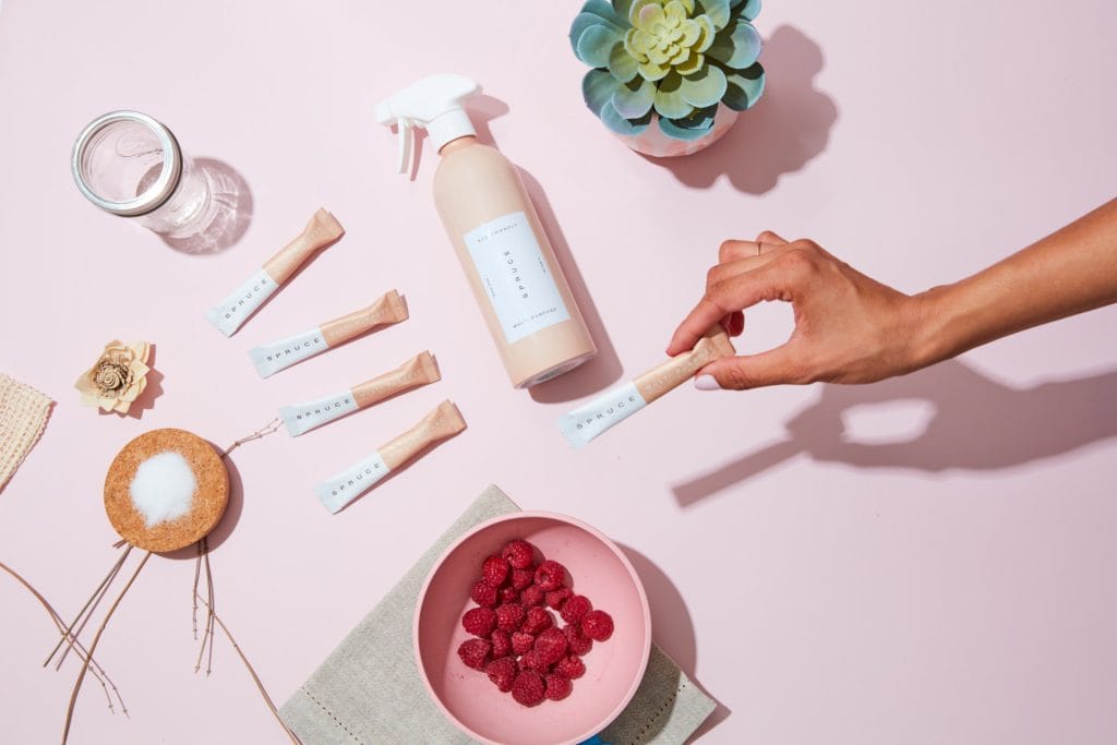 A hand picks up a small tube among cleaning product items, a spray bottle, a succulent plant, and more—all arranged on a pink surface, reflecting the tidy aesthetic of an eco-friendly Airbnb.
