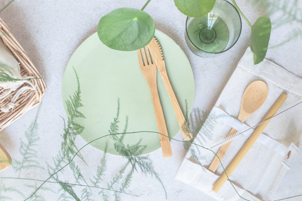 A green plate with wooden fork and knife, a glass, and a canvas pouch holding a wooden spoon and chopsticks are set on a light surface with scattered greenery—perfect for an eco-friendly Airbnb vibe.