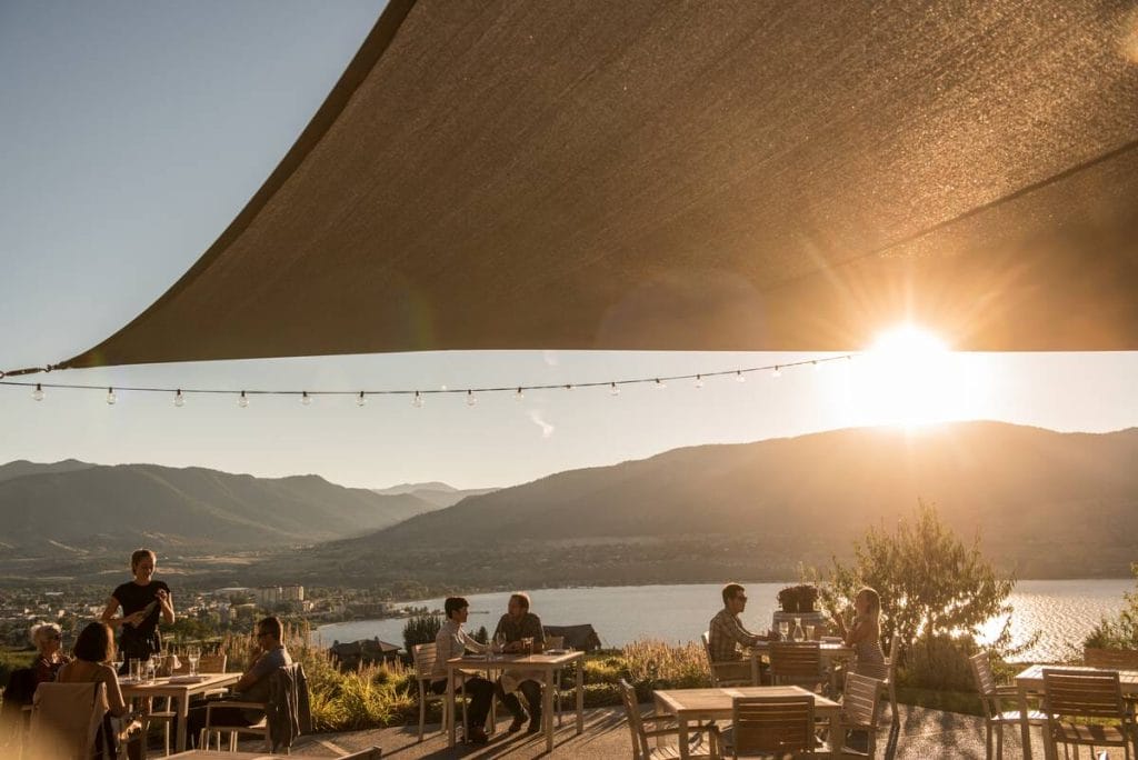 People are dining at outdoor tables under a large sunshade with string lights, overlooking a lake and mountains at sunset—a perfect setting for remote work in BC.