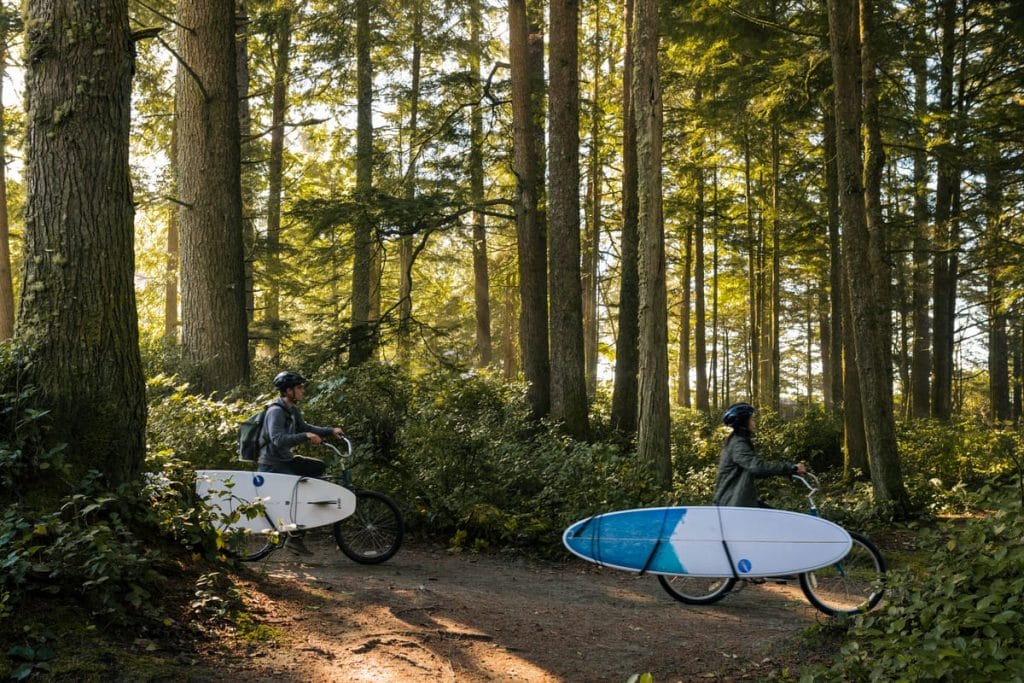 Two people ride bicycles through a forest, each carrying a surfboard attached to their bike. Sunlight filters through the trees, capturing the freedom of remote work in BC and the adventure that comes with it.