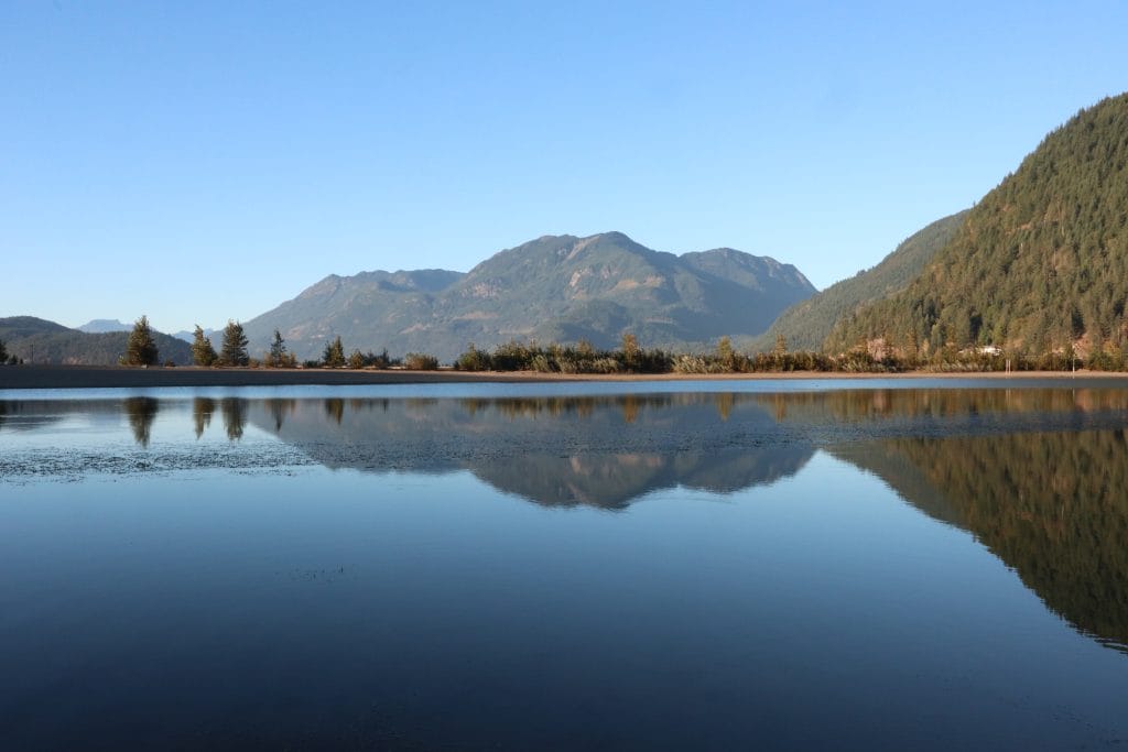 A calm lake reflects surrounding forested mountains and a clear blue sky on a bright day, evoking the tranquility that remote work in BC can offer.