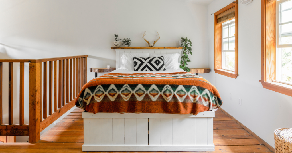 A neatly made bed with patterned pillows and a geometric blanket sits in a bright loft—an inviting example of how to setup an Airbnb, featuring wooden floors, white walls, and large windows.