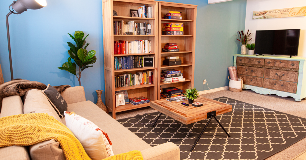 A cozy living room with a beige sofa, a wooden coffee table, a patterned rug, bookshelves filled with books and games—ideal inspiration for how to setup an Airbnb—plus a TV on a dresser and a potted plant.