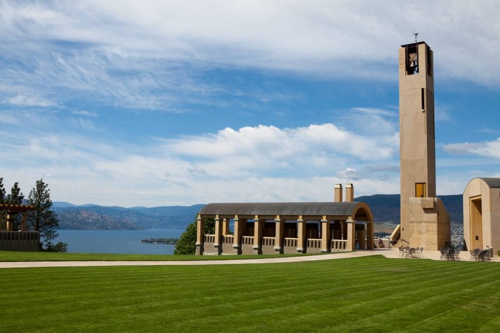 A large grassy lawn leads to a tall bell tower and arched building at one of the best wineries in West Kelowna, with a lake and mountains visible in the background under a partly cloudy sky.