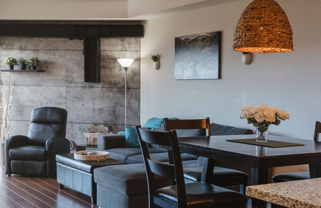 Modern living and dining area with a grey sectional sofa, recliner, dining table, wicker pendant light, and wall art. A floor lamp and potted plants decorate the space—perfect for relaxing after visiting the best wineries in West Kelowna.