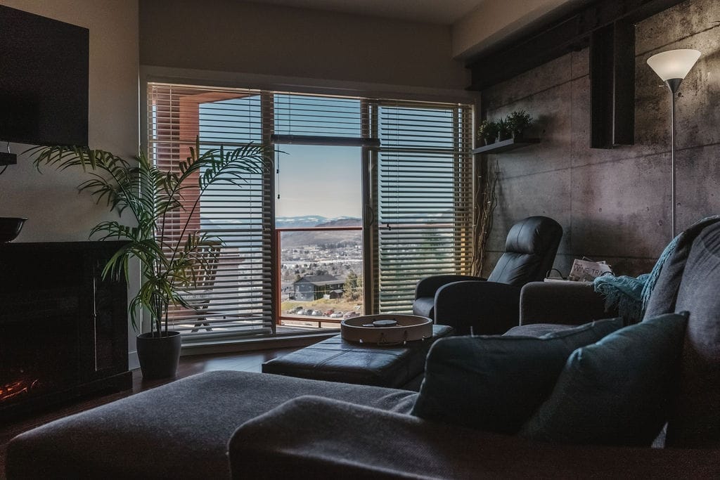 A modern living room with dark furniture, a floor lamp, a potted plant, and large windows with blinds overlooking a cityscape and mountains—perfect after a day spent exploring the best wineries in West Kelowna.