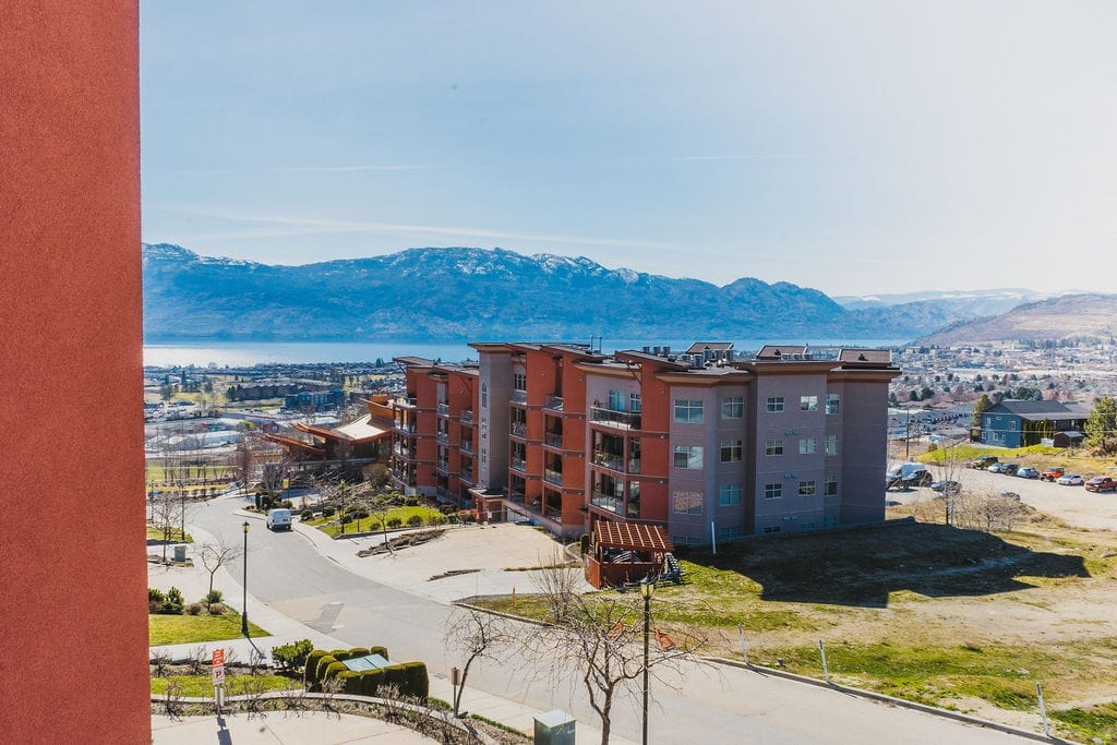 A multi-story apartment building with balconies stands near a road, overlooking mountains, a large body of water, and some of the best wineries in West Kelowna, all beneath a clear sky.