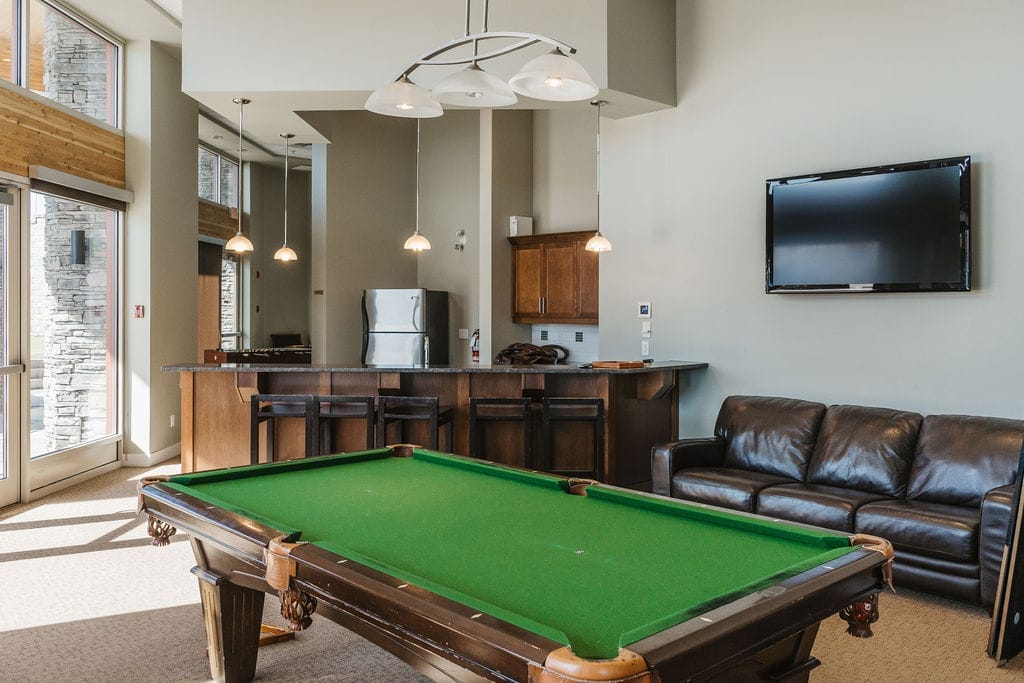 A modern lounge area with a pool table, brown leather sofa, wall-mounted TV, and a kitchen with stainless steel appliances—perfect for relaxing after visiting the best wineries in West Kelowna.