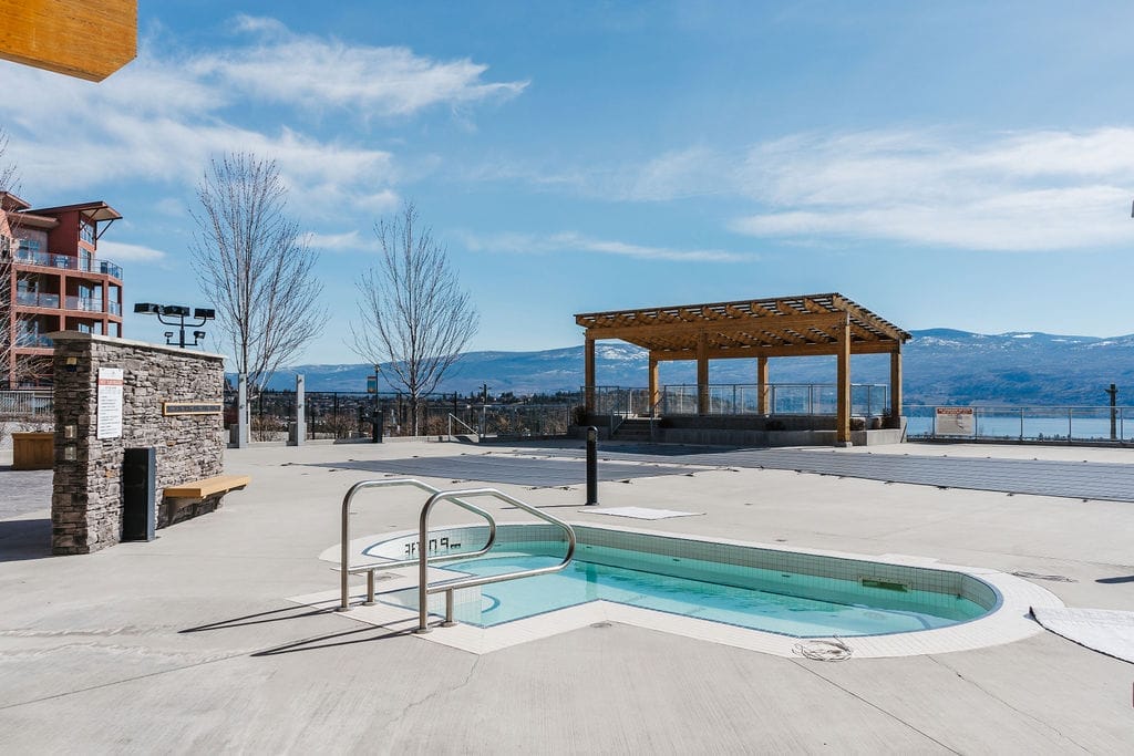 Outdoor hot tub with metal handrails on a concrete patio, a wooden pergola nearby, and mountains visible in the background under a clear blue sky—perfect for relaxing after exploring the best wineries in West Kelowna.