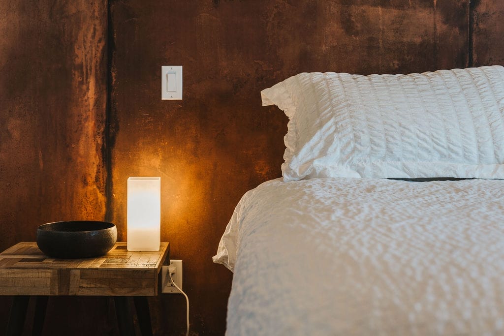 A wooden bedside table with a black bowl and a lit rectangular lamp sits next to a bed with a white textured pillow and blanket against a brown wall—perfect for relaxing after exploring the best wineries in West Kelowna.