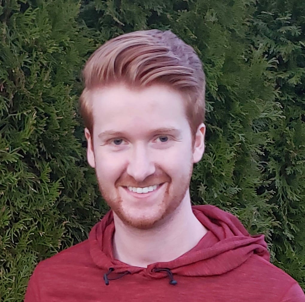 A young man with short, styled red hair and a beard smiles at the camera, wearing a maroon Lifty Life hoodie, standing in front of green foliage.